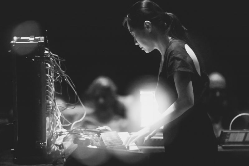 In black and white, Monica Lim concentrates on a keyboard attached with multiple wires, an audience dimly visible in the background