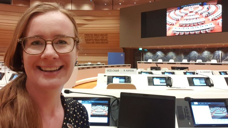 Philippa Duell-Piening beams at the camera. Behind her is a round conference room similar to a United Nations assembly, with microphones and monitors at each chair