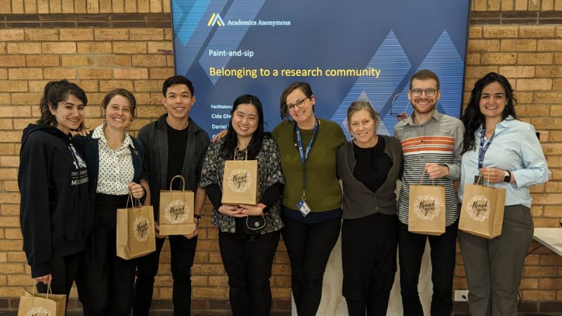Cida Ghosn beams at the camera wth a group of people of various genders and ethnicities, in front of a sign for an Academic Anonymous paint and sip event titled Belonging to a research community