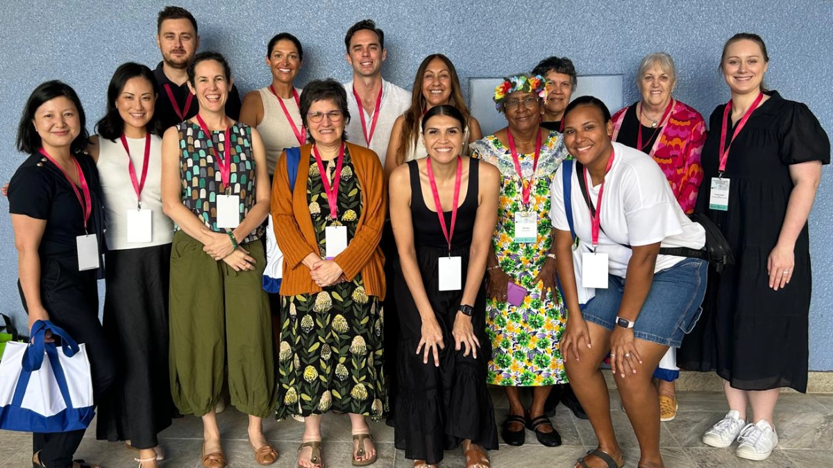 Jesse Zanker poses in a group of people of various ethnicities, ages and genders, though the group appears to be predominantly women. All wear a pink conference lanyard and are dressed for warm weather.