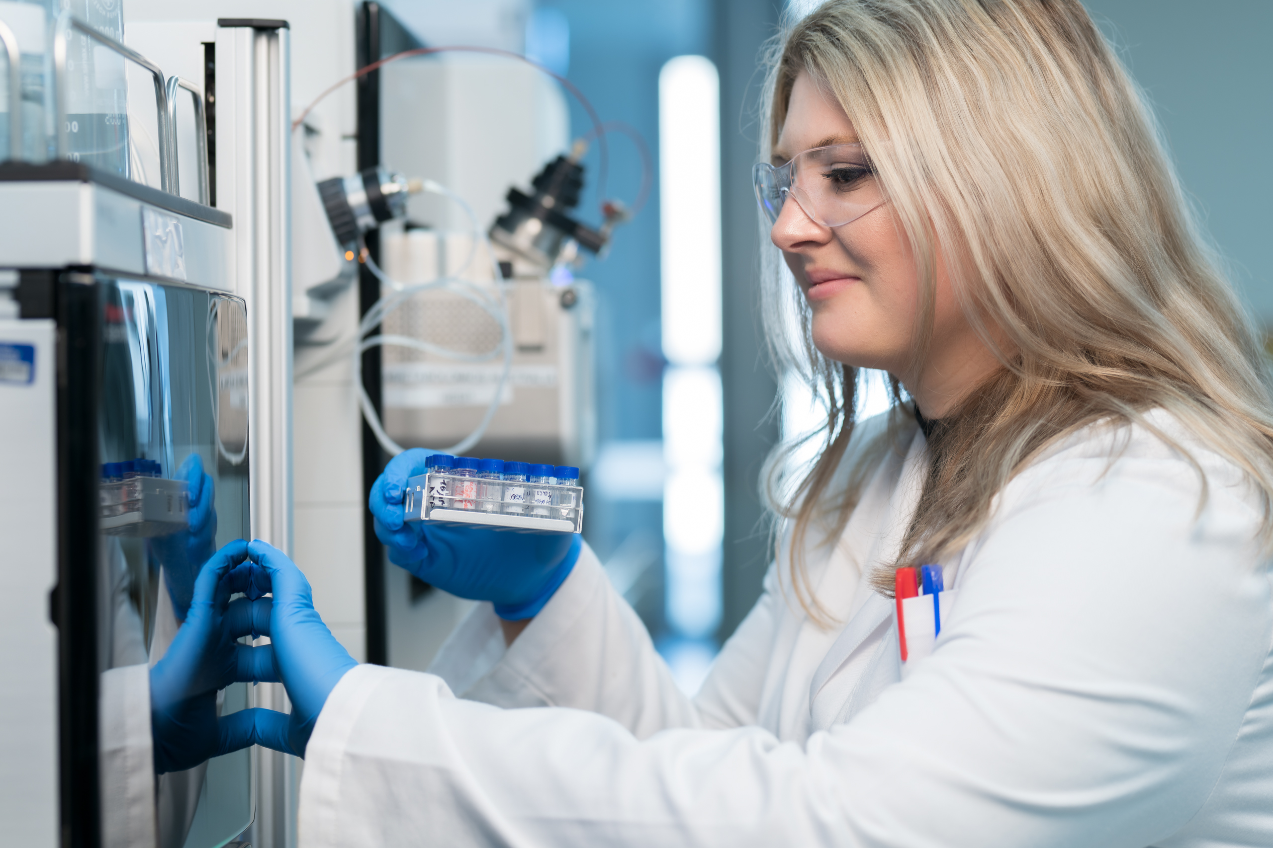 A scientist wearing a white lab coat and safety glasses works with a lab instrument.