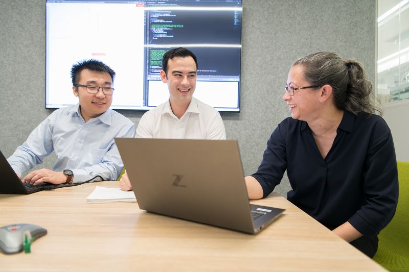 Three people smiling and collaborating at a conference table with laptops. 