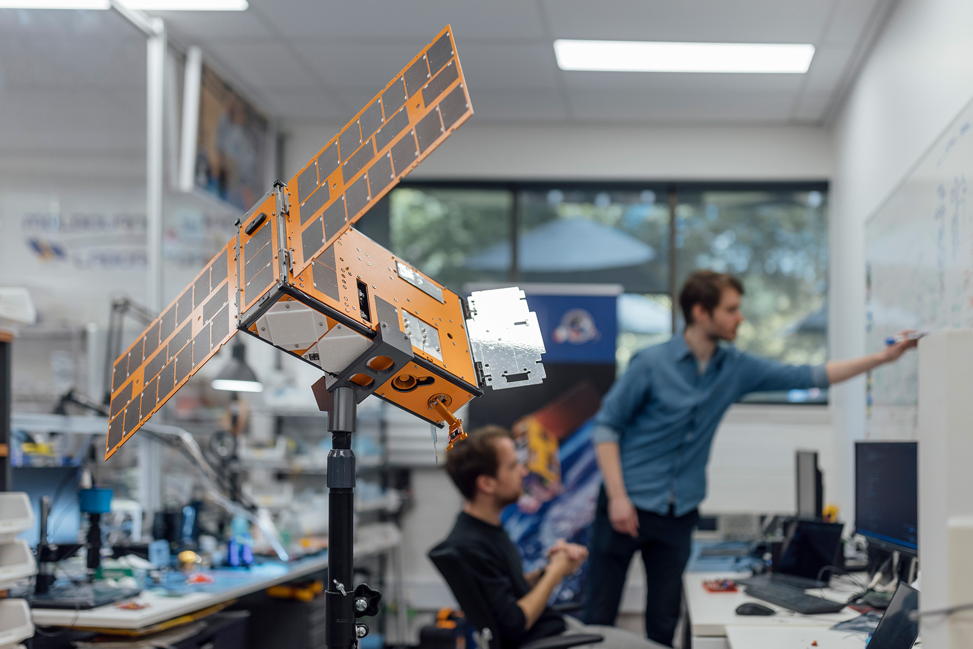 Small orange satellite in a lab with engineers brainstorming in the background