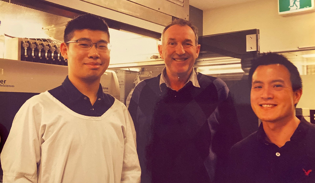 A younger Shunfei Yan wearing protective lab clothing with Rick Pearson, an older white man, and a third person, posing together in a laboratory