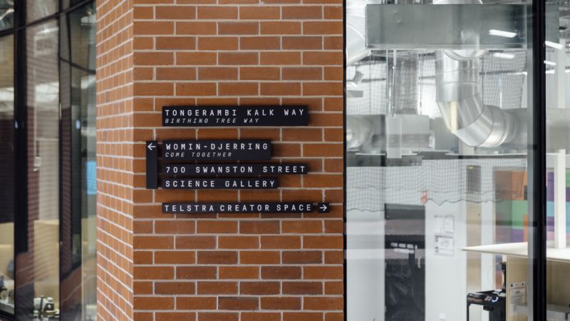 On a brick wall, signs point to the Science Gallery at 700 Swanston Street, as well as Telstra Creator Space. Machines and ducting from the Creator Space are visible through a window