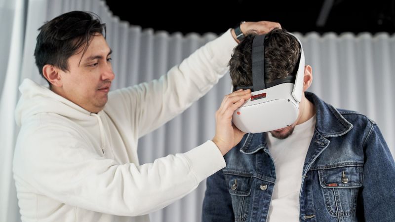Martin Reinoso, a Hispanic man with short black hair, fits a virtual reality headset on another person’s head.
