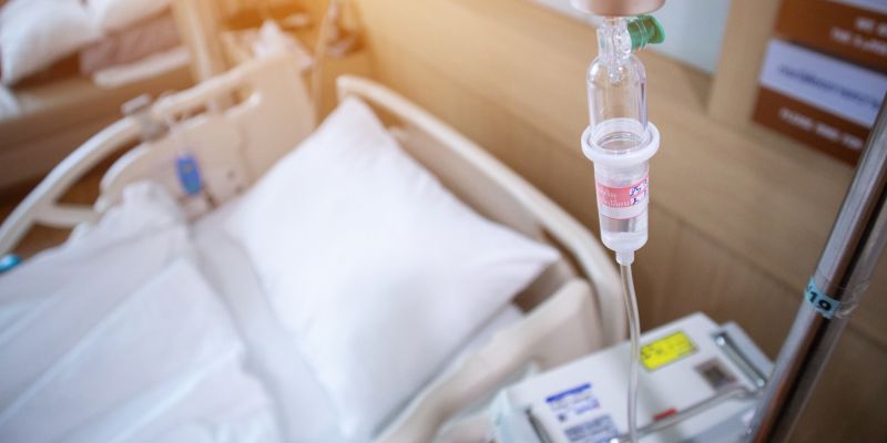 An empty, neatly made hospital bed with an IV drip in focus
