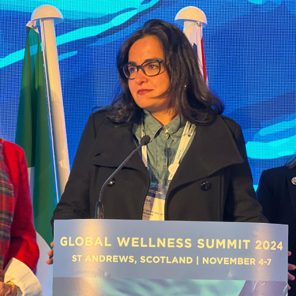 Cida Ghosn is a Brazillian woman with brown skin and long black hair. She wears glasses and a black wool jacket while speaking at the Global Wellness Summit 2024 in St Andrews, Scotland over November 4-7 