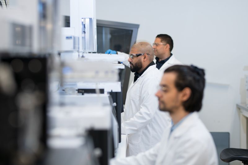Three scientists in white lab coats work in a modern laboratory, focused on equipment.