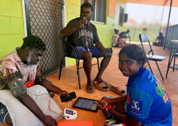 Ruth Gulamanda, Senior Yolŋu Researcher, Peter Doherty Institute, describes the photo as follows: “This photo has been taken at Djari (Rainbow) street in Galwin’ku community on Elcho Island in Arnhem Land (Yolŋu traditional lands). We went to see our [research] participant, Alvin Balwutjmi, for his health check, and these are the two workers, Fabian Malandhula and Mariana Yalumbuwuy, who work for Walking Together. The staff deliver a service outside in the community where we approach gently and sit down with them. These people are doing a Point of Care Health check for this ḏirramu (man) and he is there relaxing. This is how we do it in the community. It’s not like at the clinic, it’s [this program] outreach in the community and that’s what the Yolŋu people want – for us to go and see the people and support them with their health at their wäŋa (home). After the check we share the results with the participant, and we let them know the story the results tell. Then we do health education dhäwu (discussion) with them in Yolŋu matha (Yolŋu language) to help them understand their results and what they can do to walk on a healthy dhukarr (pathway). We enjoy our work, working with the young workers and participants in the community. The balanda (non-Indigenous) support workers help us to deliver the program. This program is good because people want to be checked at their home where they feel safe and comfortable”.