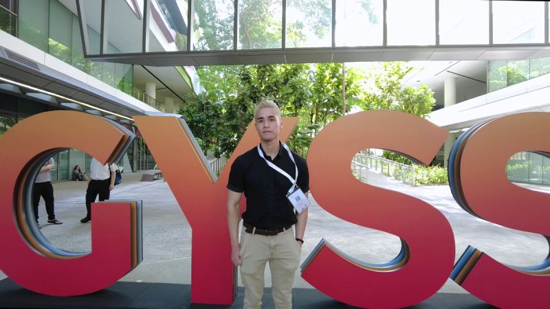 Vincent Liu poses in front of a large sign that spells GYSS