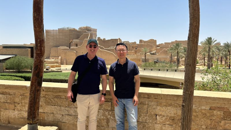 Mark Allan and a colleague pose in front of sandstone buildings in Riyadh under a blazing blue sky