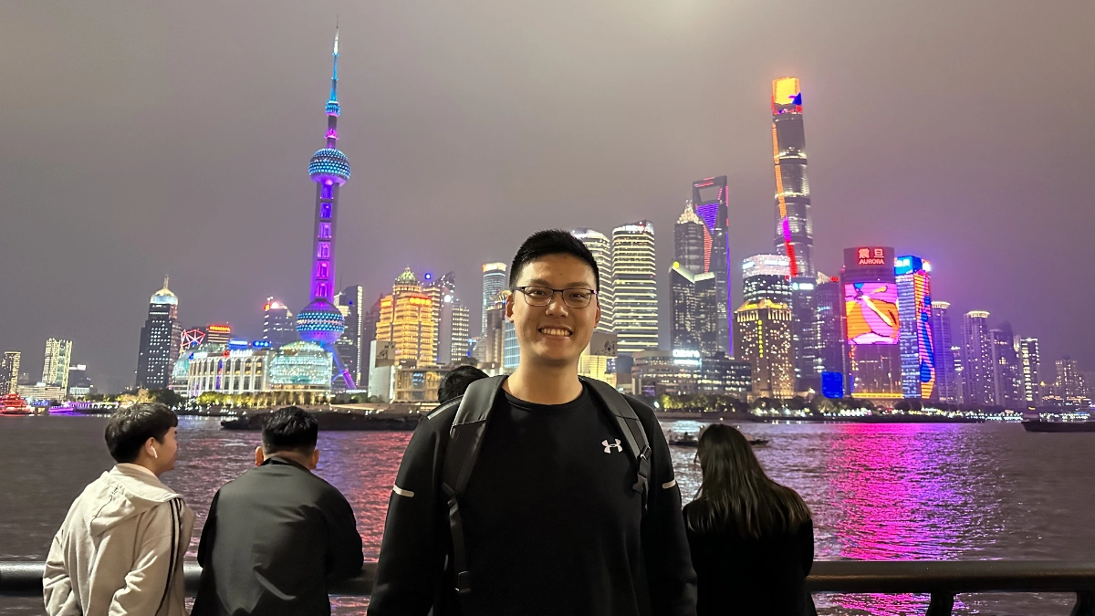 Harvey Huang is an Asian man with square thin-rimmed glasses and short black hair. He stands across the water from Shanghai's iconic skyline, lit in neon colours in the dark.