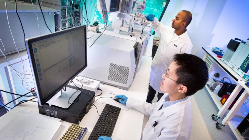 Two men in a lab wearing white lab coats. One man is using a computer, the other is checking a machine.