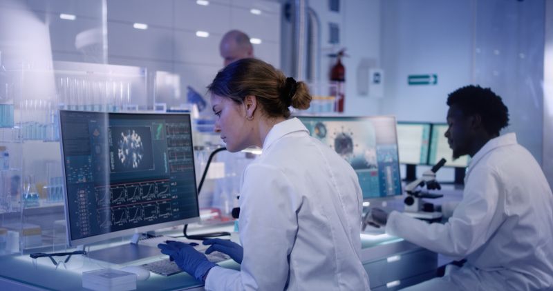 Two people in white lab coats look at screen in lab
