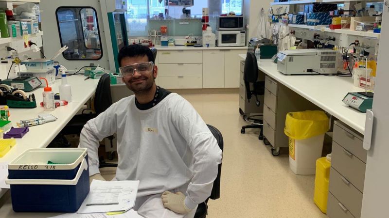 Tejas Menon, a brown-skinned man with short black hair and in a lab coat, sits at a cluttered lab space wearing protective clothing