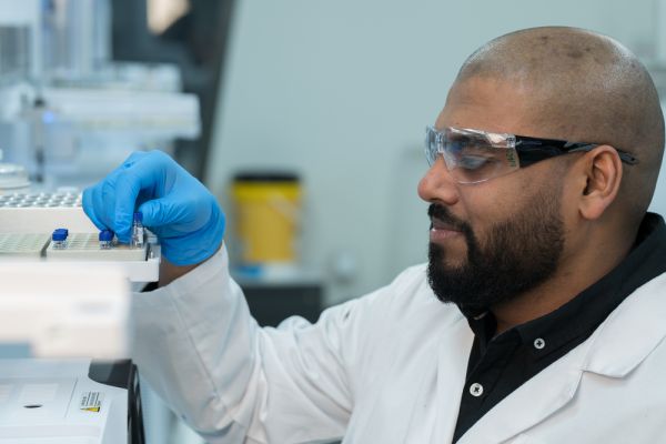 A scientist wearing safety glasses and blue gloves places a vial into laboratory equipment in a lab setting.