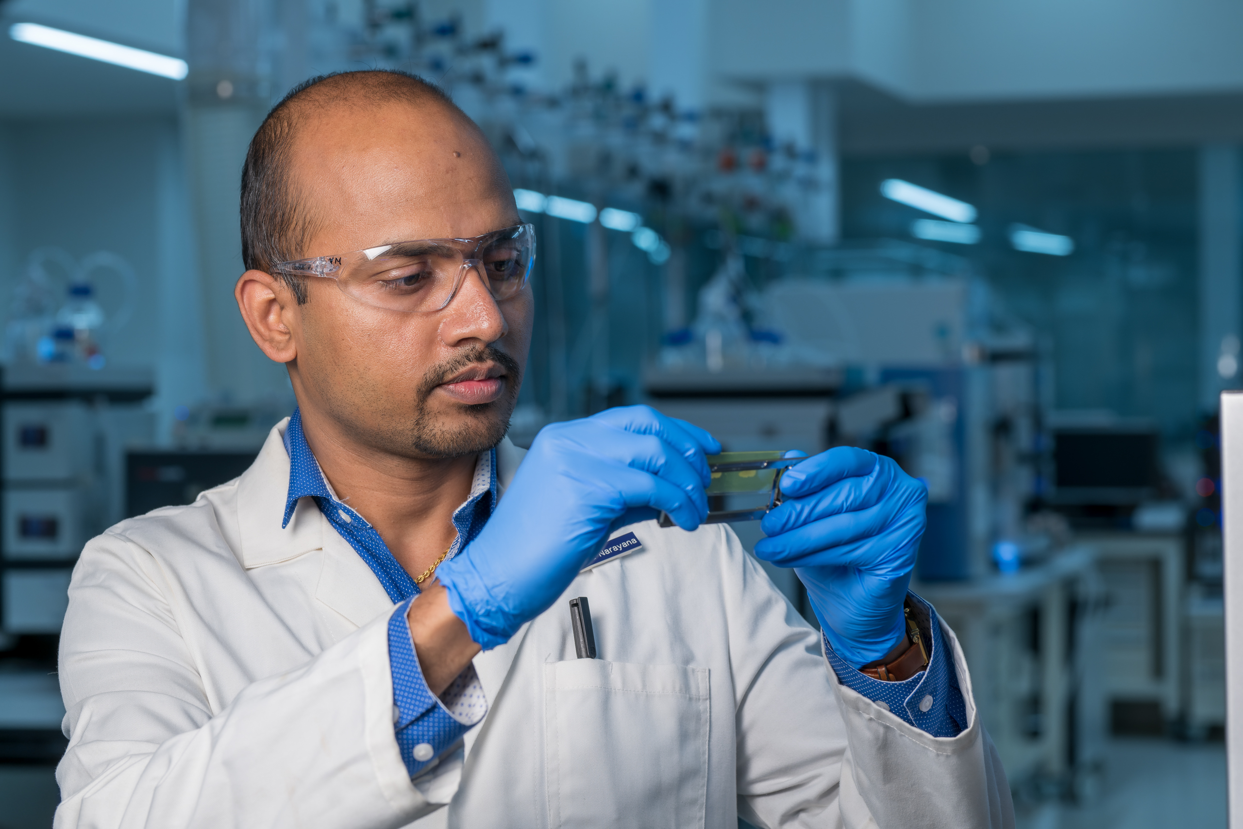 A scientist in a lab coat and goggles, wearing blue gloves, examines a slide in a modern laboratory.