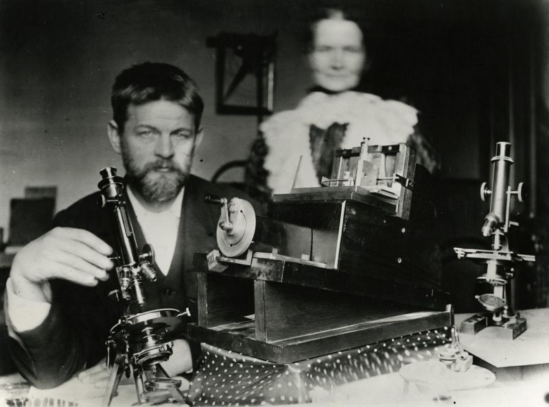Black and white photo of a man sitting at a desk, with scientific instruments. A woman stand in the background, her figure is blurred.