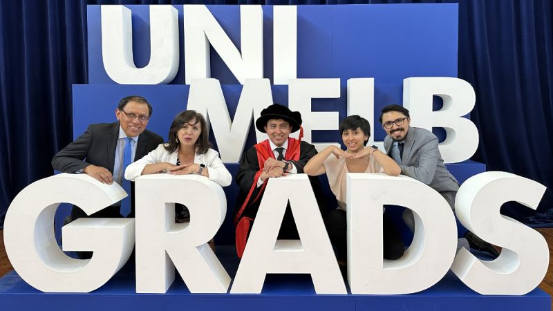 Martin Reinoso at his PhD graduation wearing academic garb for a PhD. He poses with his family behind a giant ‘UniMelb Grads’ sign.