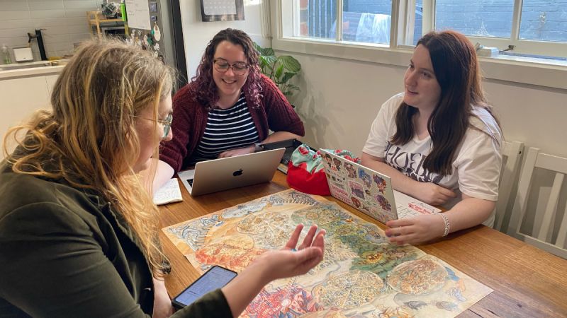 Three white women, including Nellie Seale at the head of the table, play a tabletop roleplaying game around a kitchen table, smiling.