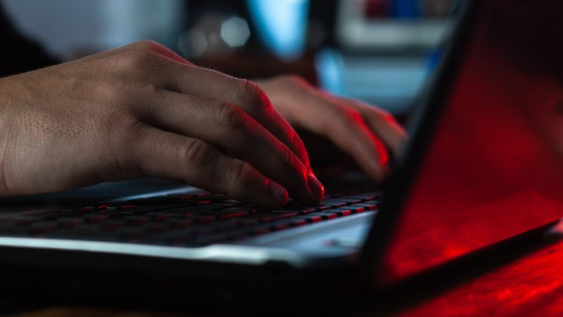 Hands type on a laptop in close focus, dimly lit by a red light