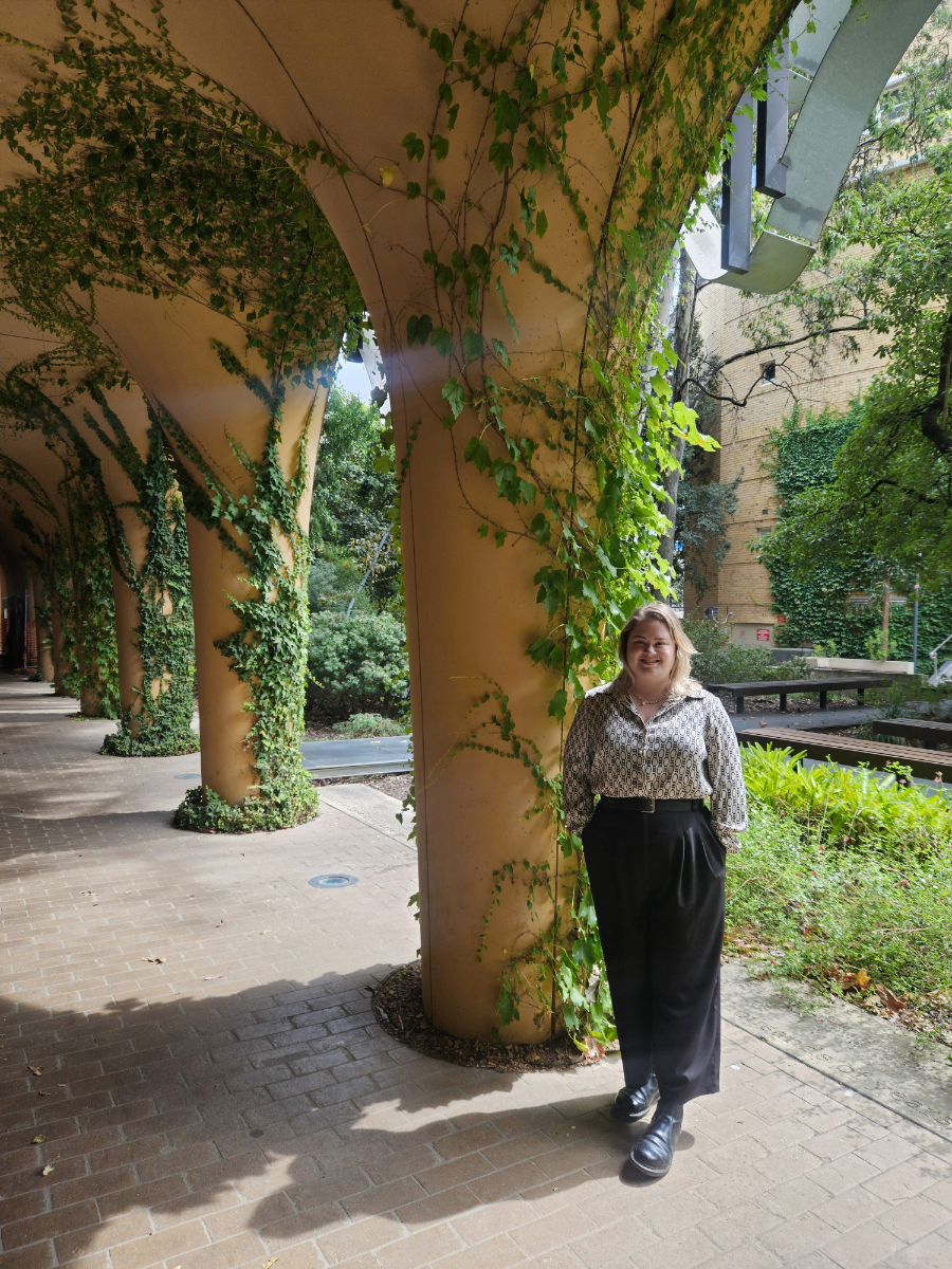 Julia Hall, a white woman, stands next to a sandstone pillar covered in vines and drenched in sunshine 