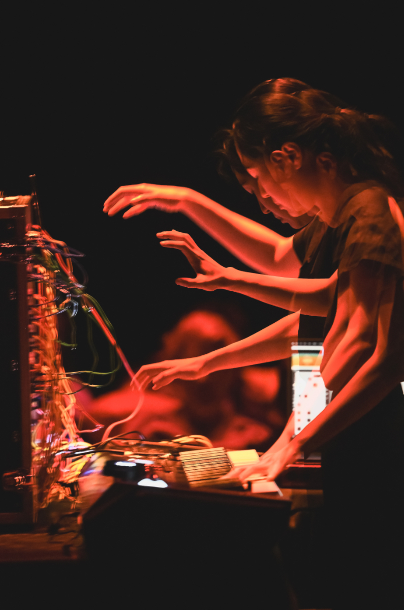 Monica Lim, an Asian woman, plays a keyboard attached with many wires, lit dimly in red. A time lapse shows two figures overlain, giving the image a sense of motion