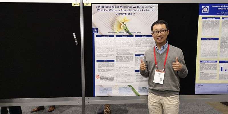 Hanchao Hou, an Asian man with short black hair, poses with thumbs up in front of a poster titled Conceptualising and Measuring Wellbeing Literacy: What Can We Learn From a Systematic Review of Literacy Studies