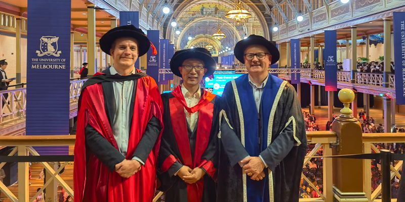 Dr Hanchao Hou wearing academic garb at his graduation in an elaborate University of Melbourne hall with his supervisor Professor Lindsay Oades, who is a white man, and a third person