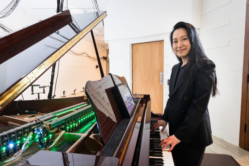 Monica Lim, an Asian woman, smiles with her fingers on the keys of a grand piano, which is lit on the inside with a row of LED lights