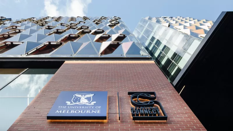 Melbourne Connect's geometric panels jut out from the facade which rises towards a blue sky. In the foreground are the University of Melbourne and Melbourne Connect's logos on a plain brick wall.