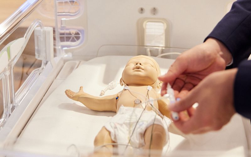 A medical baby dummy in a hospital bassinet, brown hands maneuvering lines connected to it