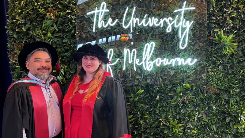 Dr Kayla Heffernan poses with her supervisor Professor Shanton Chang in front of a University of Melbourne sign, both wearing academic dress for PhDs. 