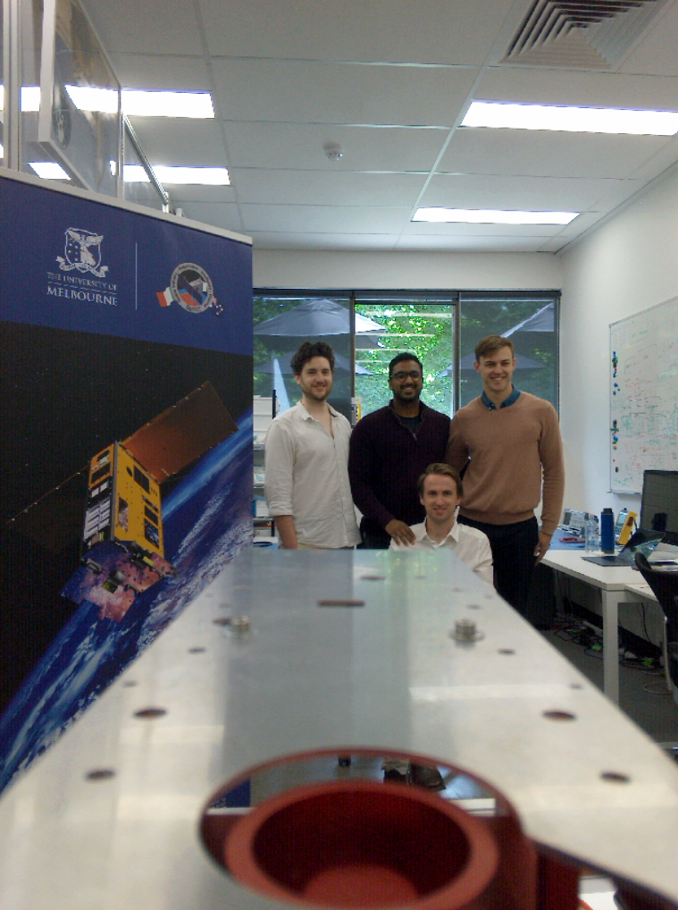 Four members of the Melbourne Space Lab next to a banner featuring the SpIRIT nanosatelliterne Space Lab stand next to a