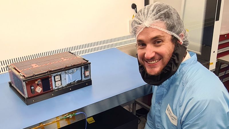 Rob Mearns, a white man, wears protective clothing and smiles broadly next to a shoebox-sized satellite