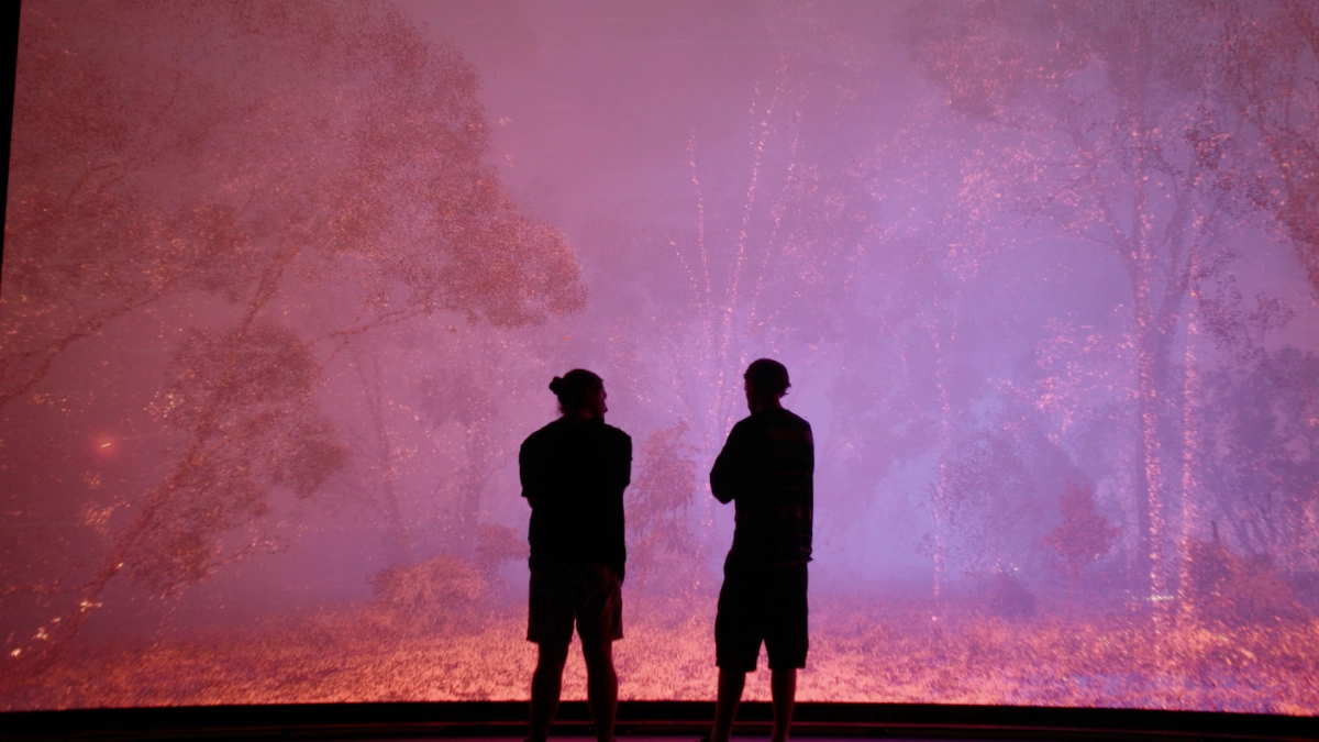 Two people are silhouetted against a giant screen depicting what looks like a bushfire, lit yellow and red with a purple smoke obscuring the scene