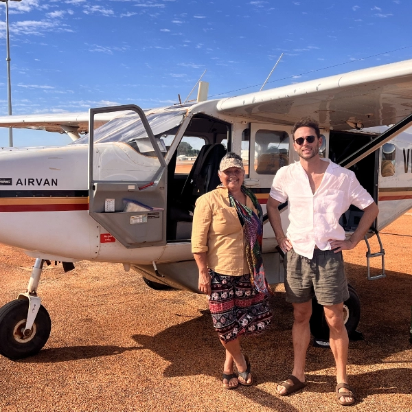 Jesse Zanker is a young white man dressed in linen shorts, a white shirt and sandals. He smiles next to Aunty Ros Malay, an Indigenous woman in a linen shirt, flowy patterned skirt and sandals. They stand in front of a small aircraft on a rusty red airfield, blue skies abovehead.