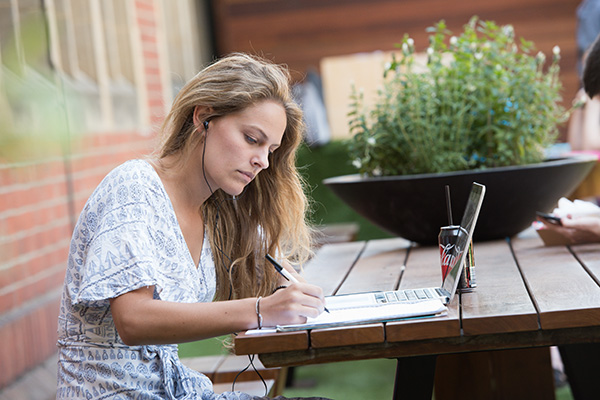 A light-skinned woman with long blonde hair writes in a notebook on a picnic table outdoors