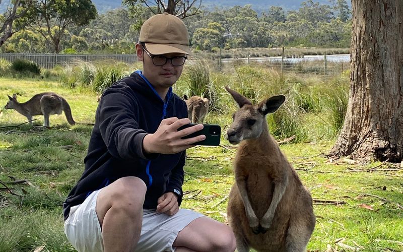 Dr John Lu, an Asian man wearing glasses, a cap and comfortable clothes, poses next to a wallaby on a grassy background