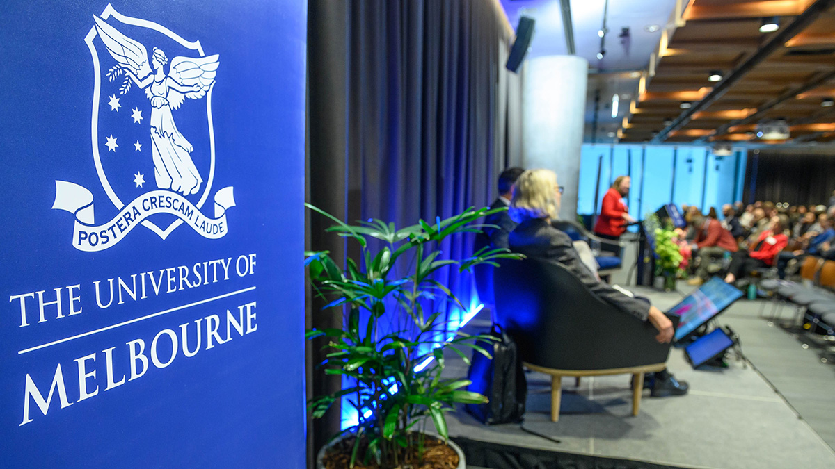 The University of Melbourne logo prominent in the foreground with the audience listening to a panel discussion in the background