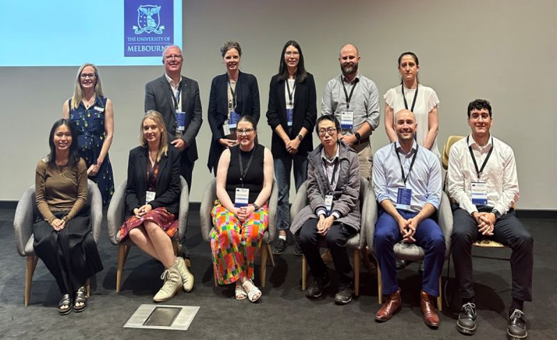 Six graduate researchers sit in a row on a stage under a University of Melbourne logo wearing business casual clothing and lanyards for a conference