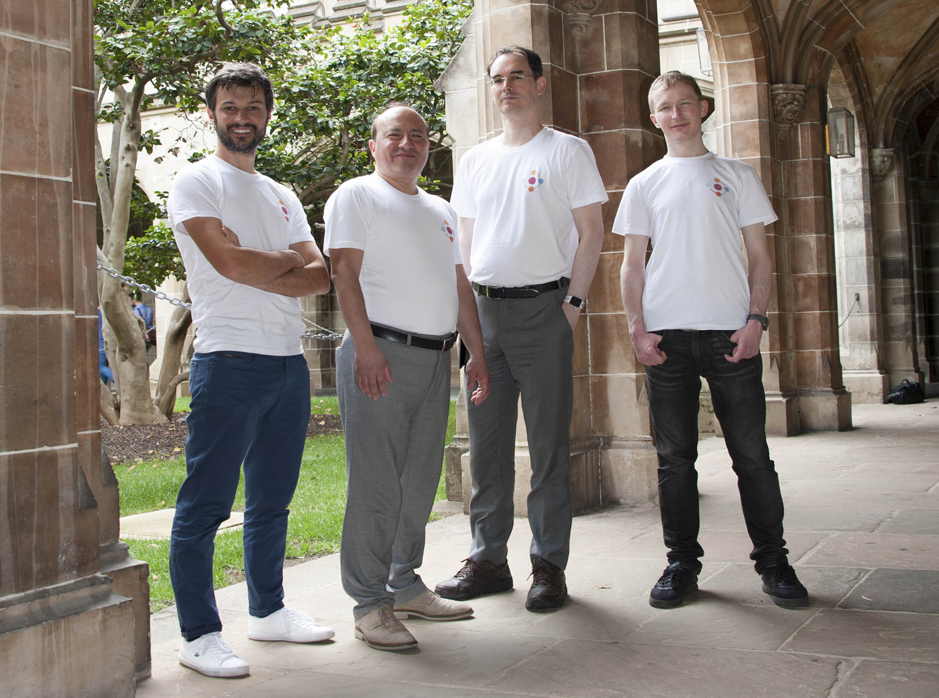 Four men in matching white t-shirts stand under a sandstone archway
